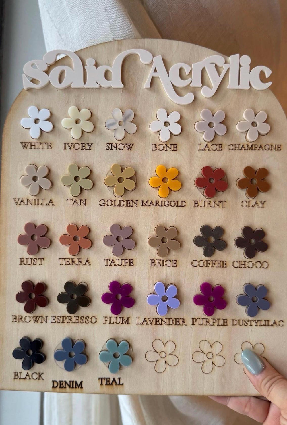 A wooden display board featuring a variety of solid acrylic flower shapes in various colors. The board is labeled "Solid Acrylic" and includes color names like "White," "Ivory," "Tan," and "Teal." The