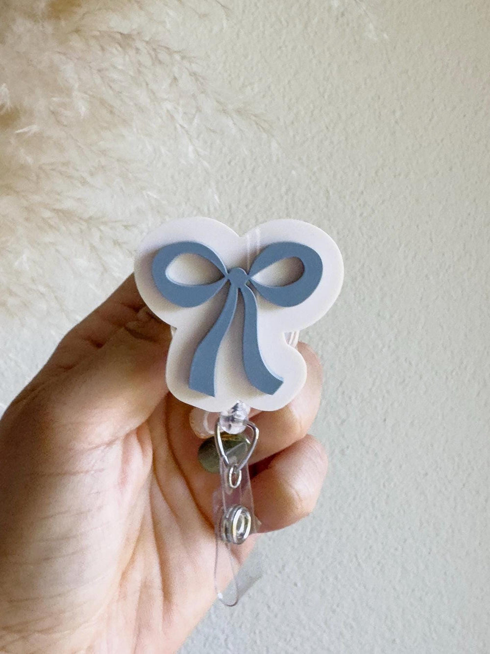 A white and blue bow-shaped badge reel. The badge reel features a light blue bow design on a white background. The reel has a clear plastic clip and a retractable cord.
