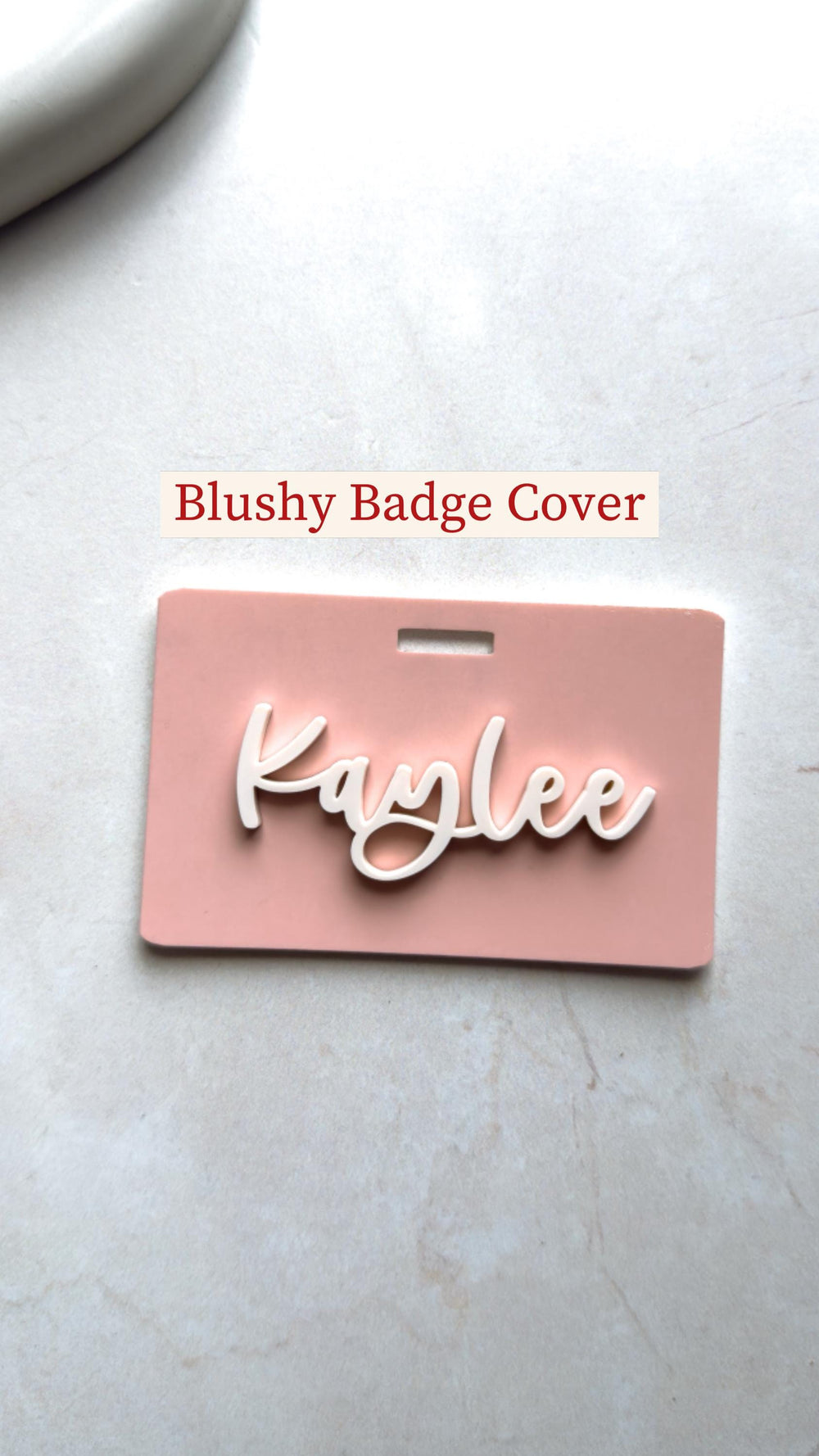 A blush pink badge cover with the name "Kaylee" in white cursive script. The badge cover has a rectangular shape with a slot at the top. The text "Blushy Badge Cover" is above the badge.