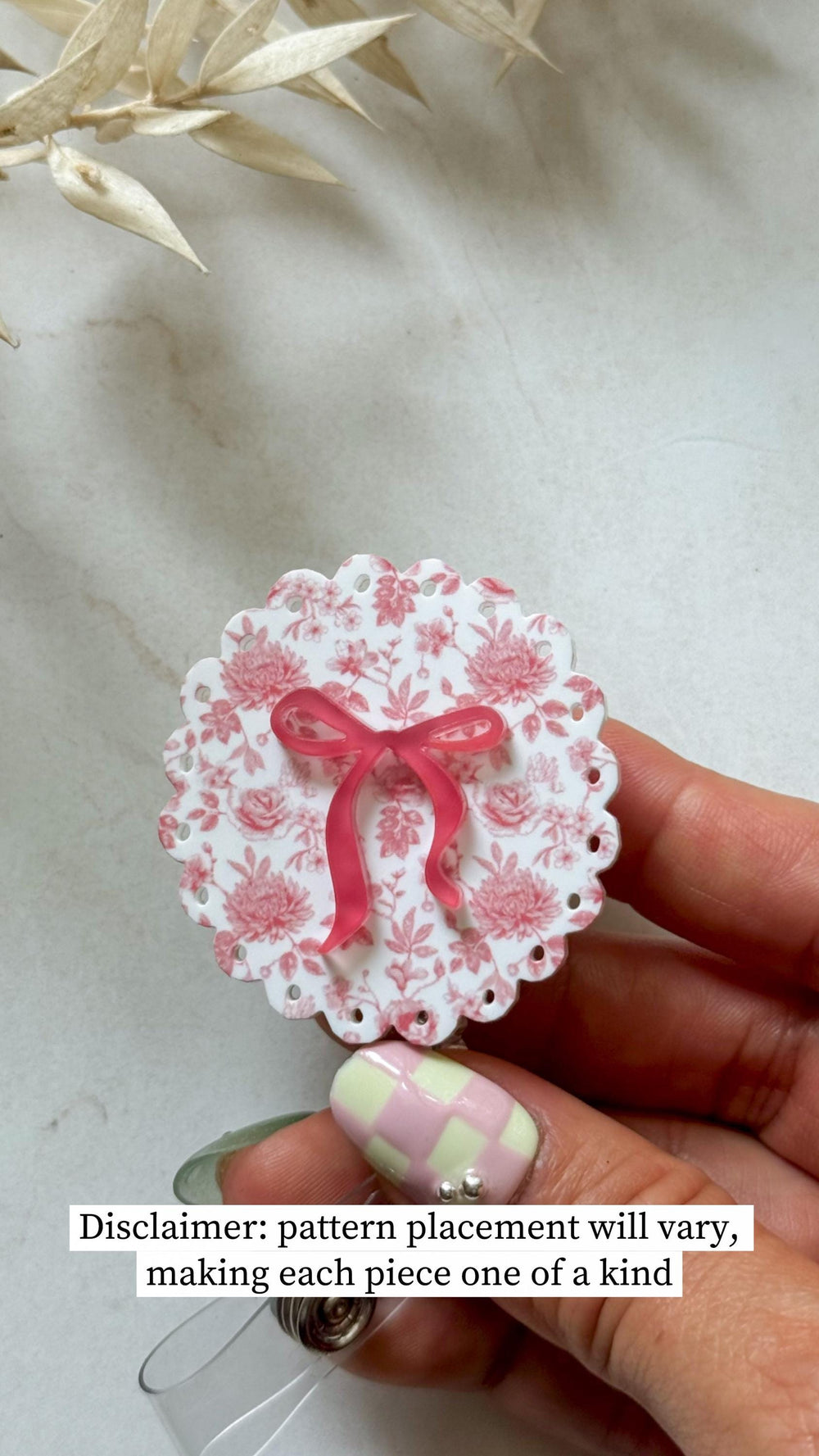 A round badge reel with a floral pattern in pink and white, topped with a pink bow. The edge is scalloped with small holes. The text reads: "Disclaimer: pattern placement will vary, making each piece 
