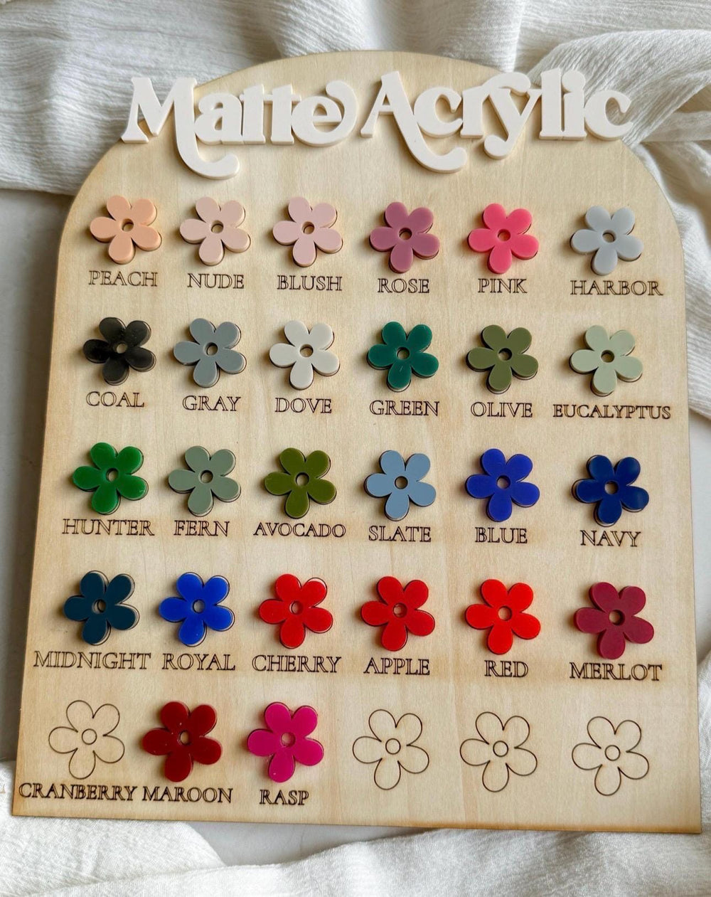 A wooden color chart with the words "Matte Acrylic" at the top. It displays an array of flower-shaped color samples in various shades, including peach, pink, blue, and red. Each color is labeled.