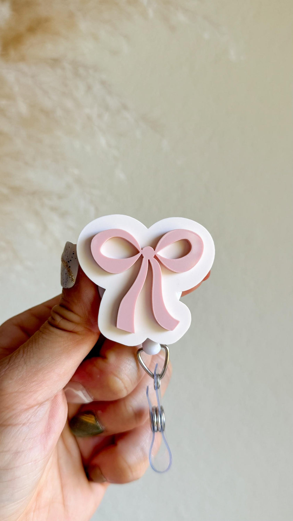 A white and pink bow-shaped badge reel. The bow is light pink and sits on a white background. The reel has a silver clip and a clear plastic cord for attaching to clothing or accessories. The design i