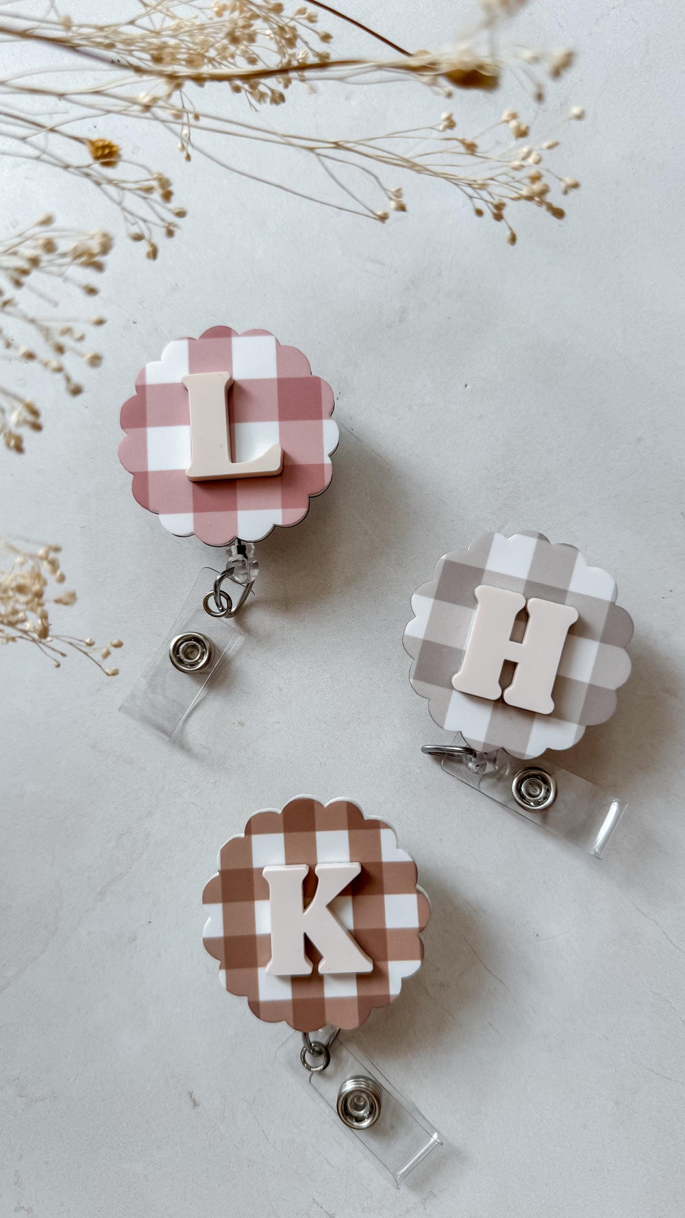Three floral-shaped badge reels with gingham patterns in pink, tan, and brown. Each reel features a different letter: L, H, and K, in a complementary color. The reels have a clear plastic strap and a 
