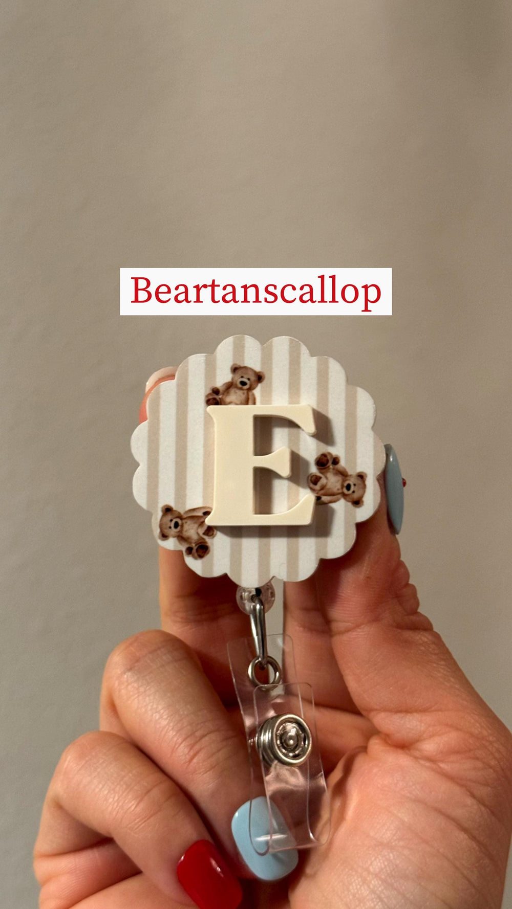 A badge reel with a scalloped edge and a beige and white striped background. The badge features a large, cream-colored letter "E" and three small teddy bear illustrations. The text "Beartanscallop" is