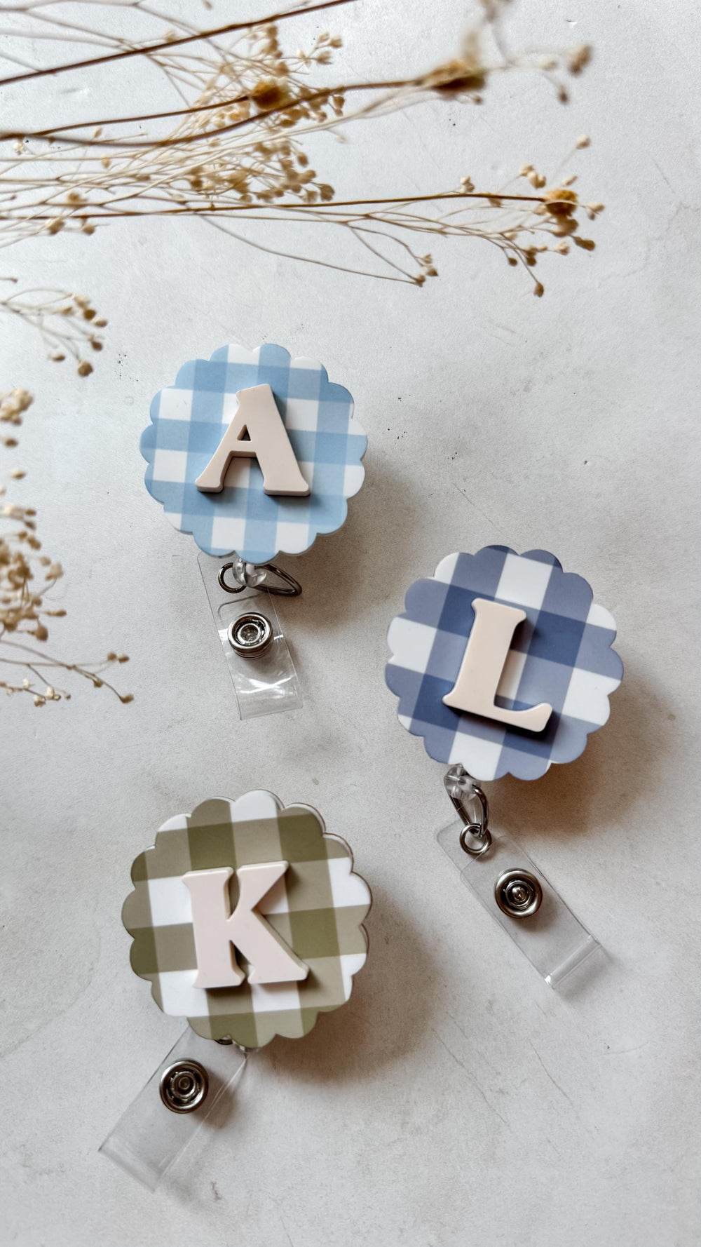 Three badge reels with scalloped edges and gingham patterns. One is light blue with the letter "A", another is blue with "L", and the third is olive green with "K". Each has a clear plastic strap and 