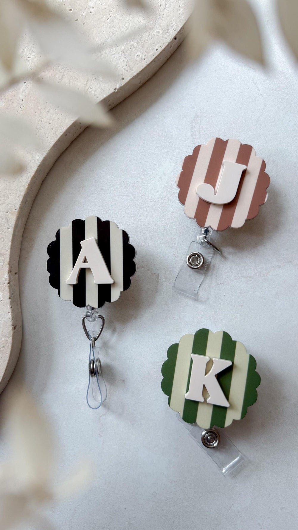 Three personalized badge reels with scalloped edges. One has black and white stripes with a white "A", another has brown and white stripes with a white "J", and the last has green and white stripes wi
