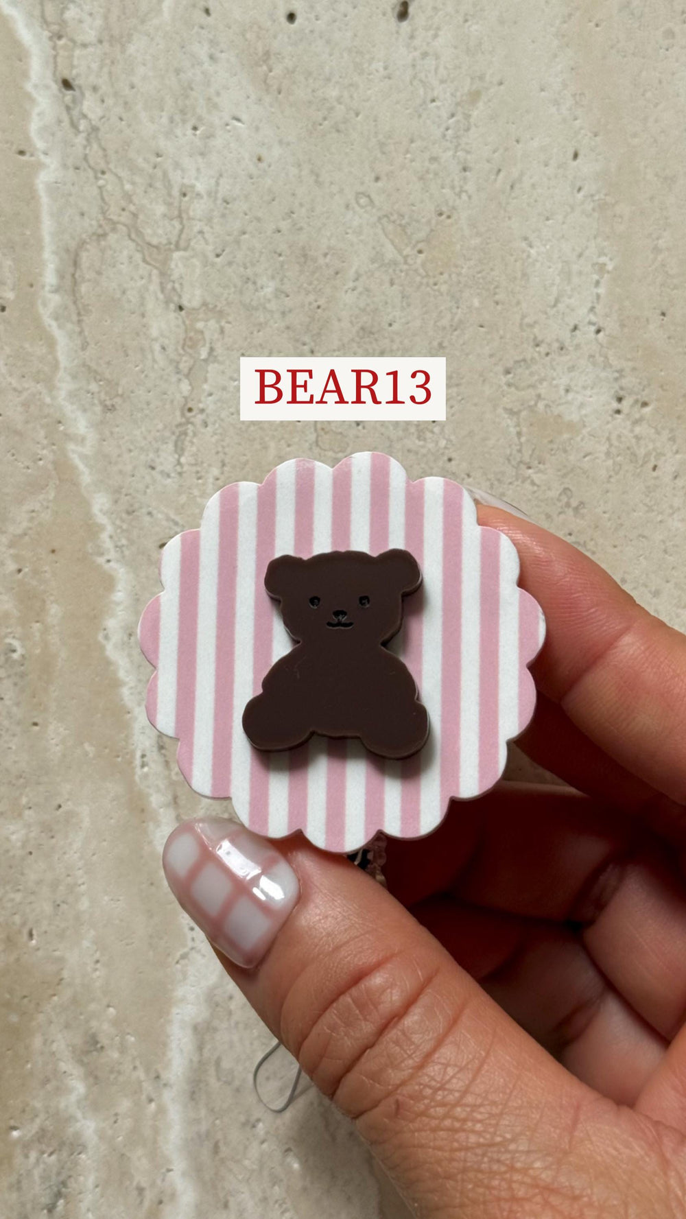 A brown teddy bear-shaped embellishment on a pink and white striped background. The embellishment is set on a white, scalloped edge. The text "BEAR13" is displayed in a white rectangle.