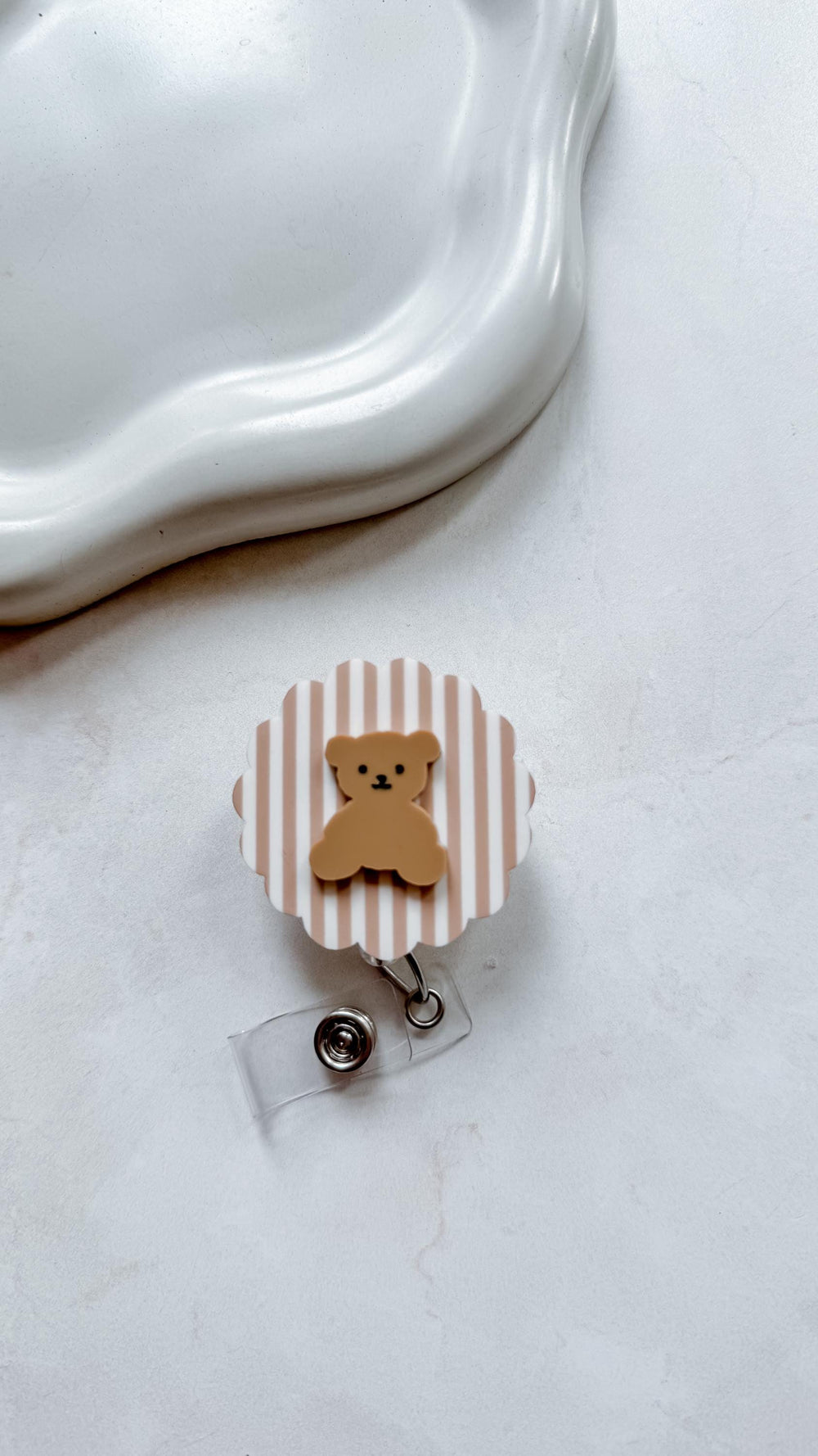 A badge reel with a brown teddy bear design. The reel has a white and tan striped background with a clear plastic clip and a metal snap closure. The badge reel is a cute accessory.