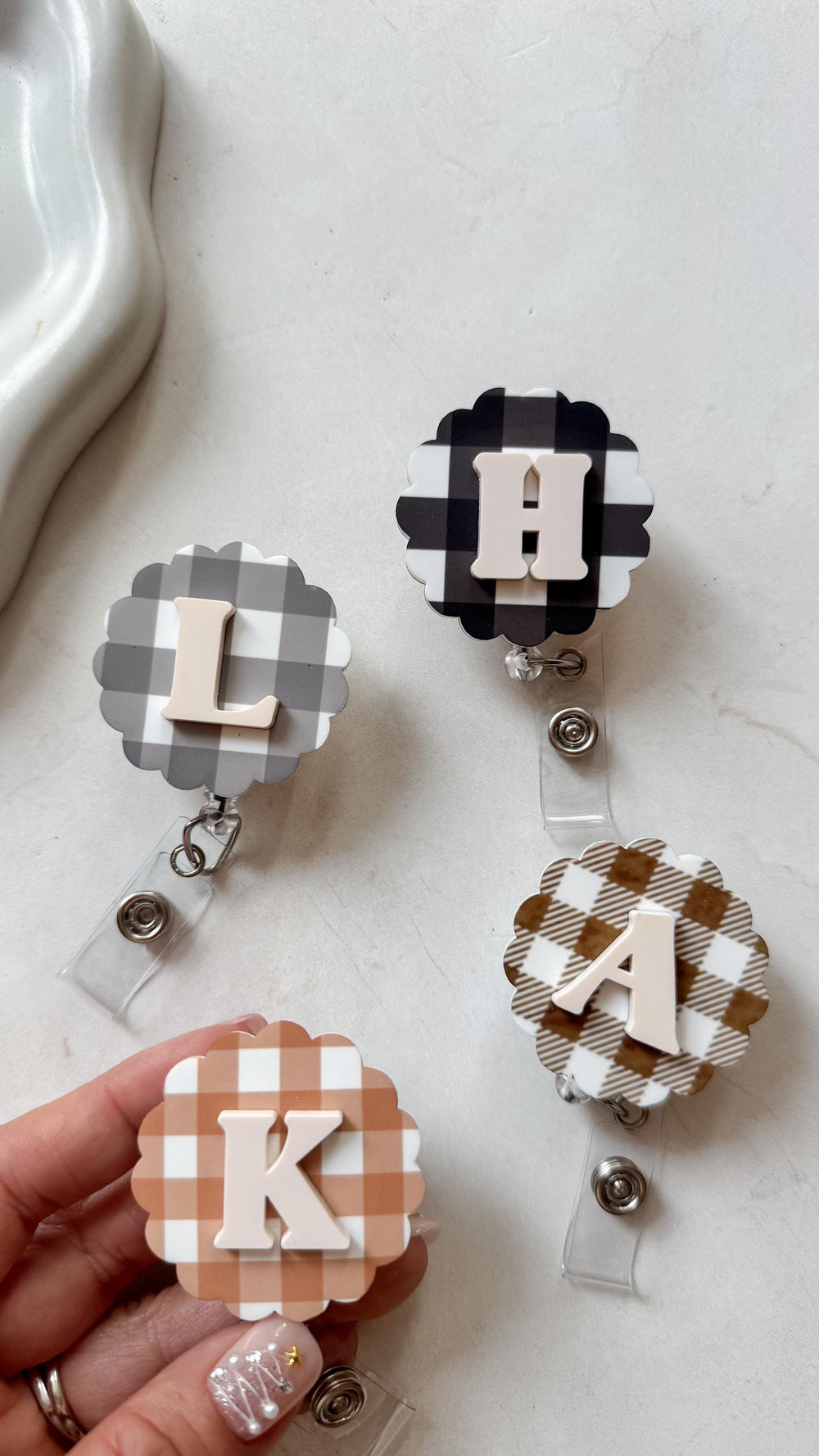 Four badge reels with scalloped edges and gingham patterns in black and white, gray, and brown. Each reel features a different letter in a neutral color: L, H, A, and K. The reels have a clear plastic
