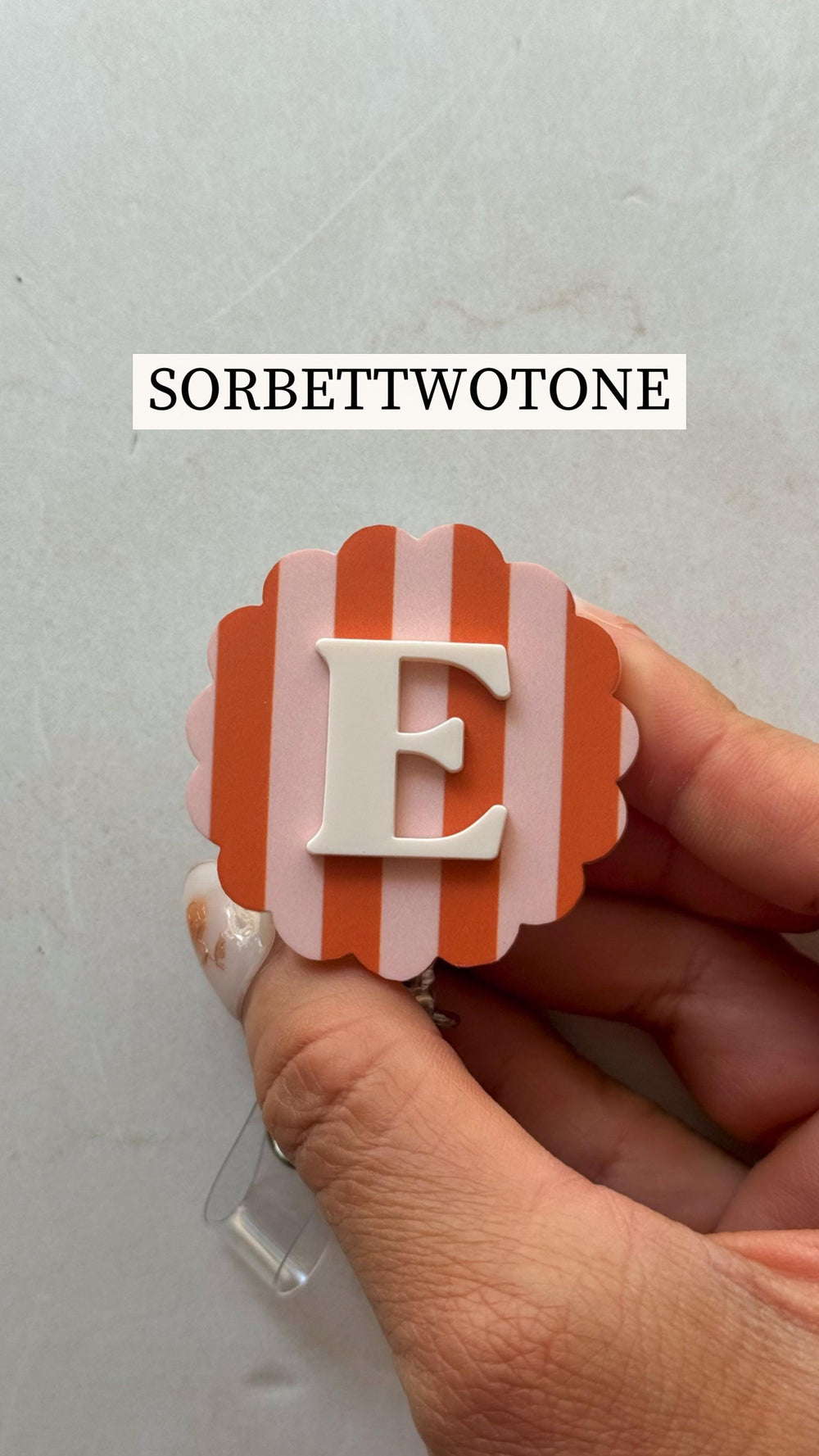 A badge reel with a scalloped edge, featuring a pink and orange striped background and a white letter "E". The text "SORBETTWOTONE" is displayed above the badge reel.